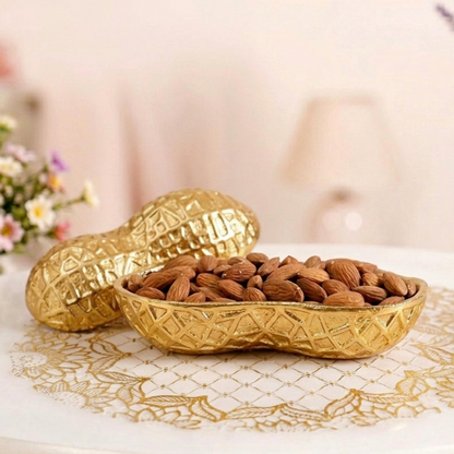 A decorative gold serving bowl shaped like a peanut shell, filled with raw almonds. The matching textured gold lid rests beside it on a white table adorned with a gold lace doily. The background is softly blurred, showing hints of floral arrangements and a warm lamp.