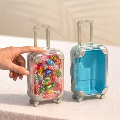A hand holding a clear pink mini suitcase organizer filled with colorful candies, placed next to an empty blue miniature travel storage box on a textured table - saathihome
