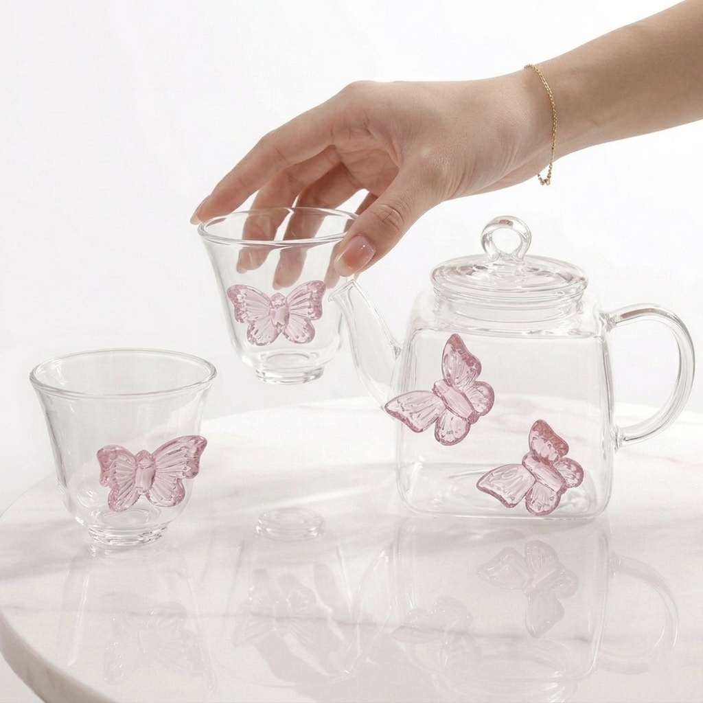 ​A woman's hand reaching to pick up a clear glass teacup featuring a 3D pink butterfly design, positioned next to a matching glass teapot on a light marble surface.
