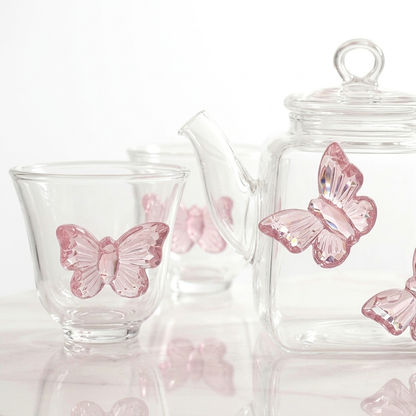 Empty transparent glass teapot and two matching cups decorated with applied pink glass butterflies, resting on a white marble tabletop in a brightly lit room.