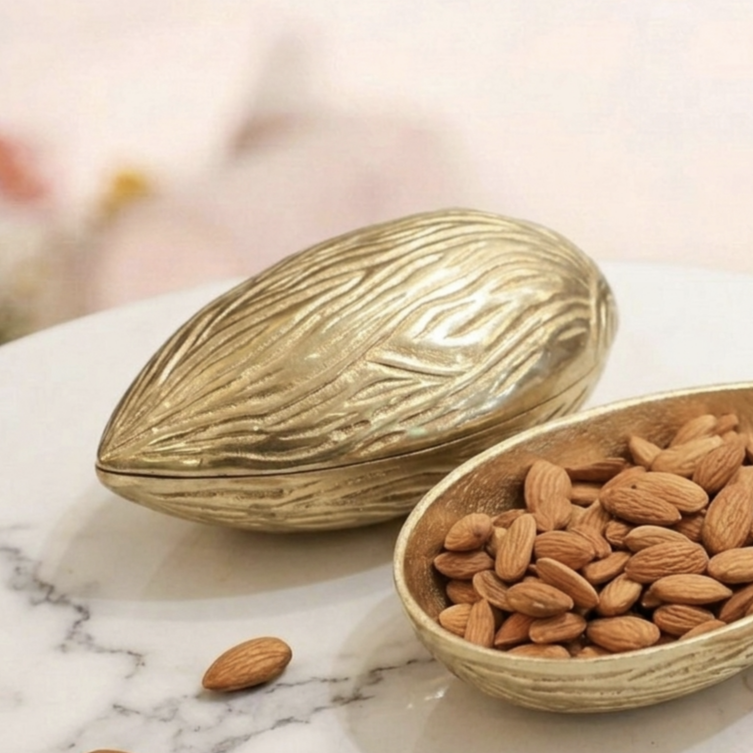 Golden brass storage bowl shaped like a textured almond, partially open with its lid resting to the side, filled with raw almonds and placed on a white marble countertop.