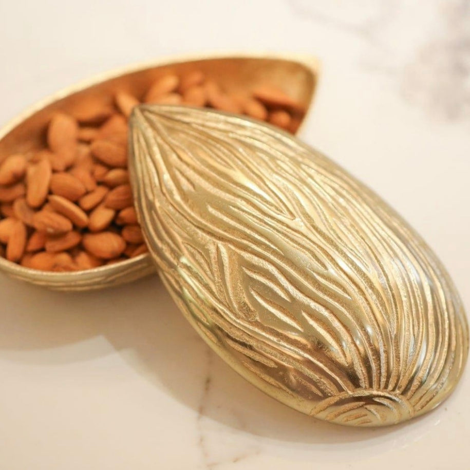 Golden brass textured storage bowl shaped like a textured almond, partially open with its lid resting to the side, filled with raw almonds and placed on a white marble countertop.