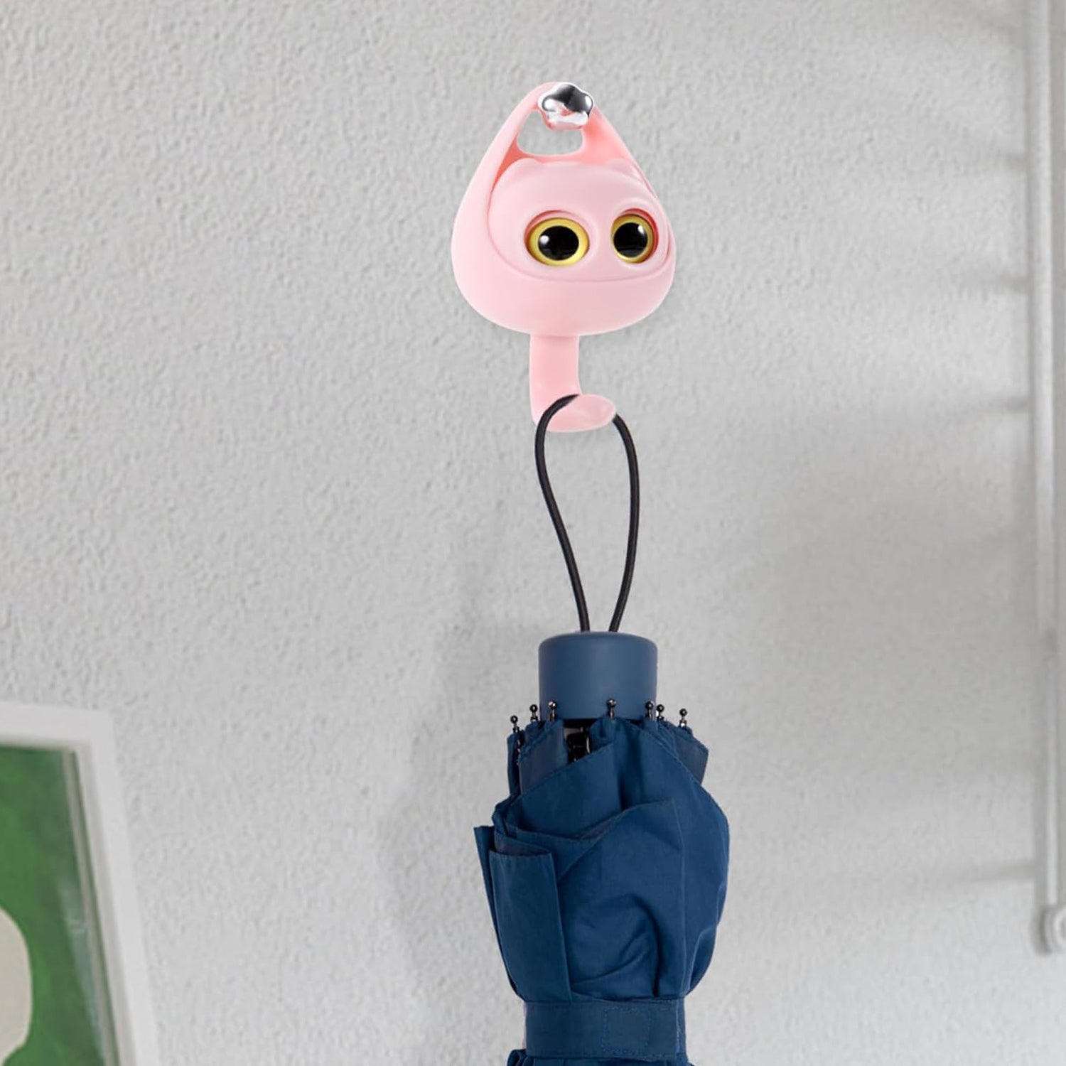 Pink cat wall hook securely holding a blue folding umbrella by its strap on a textured white wall