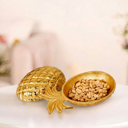 High-angle, professional product photograph of an ornate, two-piece golden pineapple-shaped server resting on a clean white pedestal. The right half of the split pineapple is a textured bowl filled with blanched peanuts, featuring a detailed golden frond leaf handle. The left half of the split pineapple is the matching textured ornate pineapple lid, resting slightly to the side. The overall presentation is against a seamless white background.